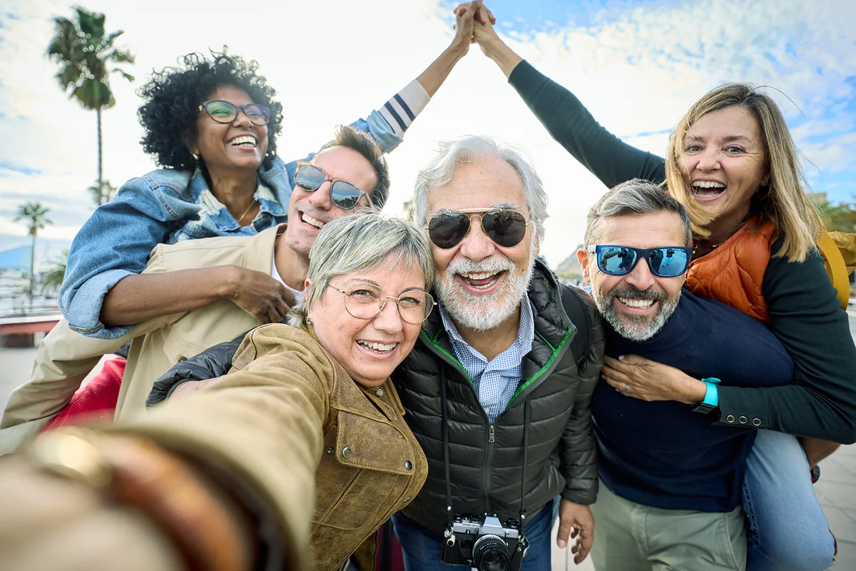 Group diverse funny mature tourist friends posing piggyback with hands joined in air taking photo selfie together with front camera on travel outdoor. Adult