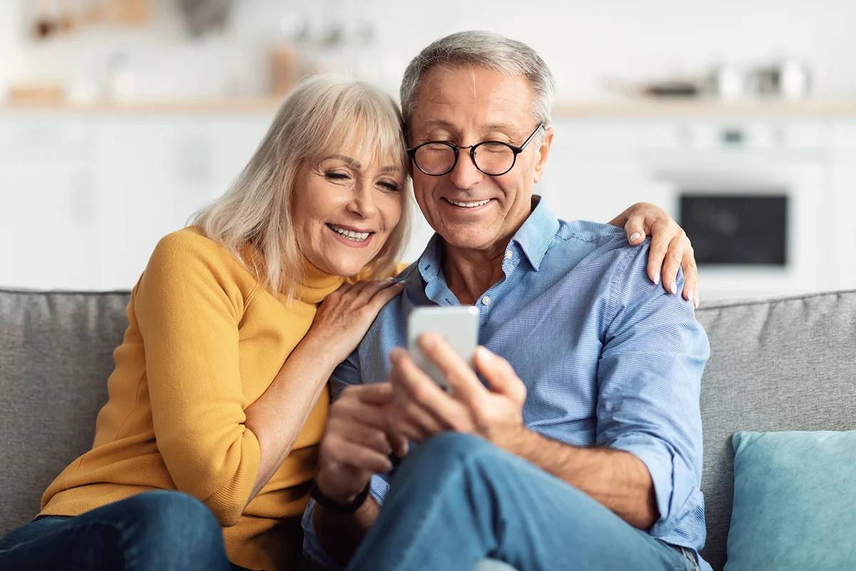Happy Senior Couple Using Smartphone Browsing Internet Together Sitting On Couch At Home And Hugging