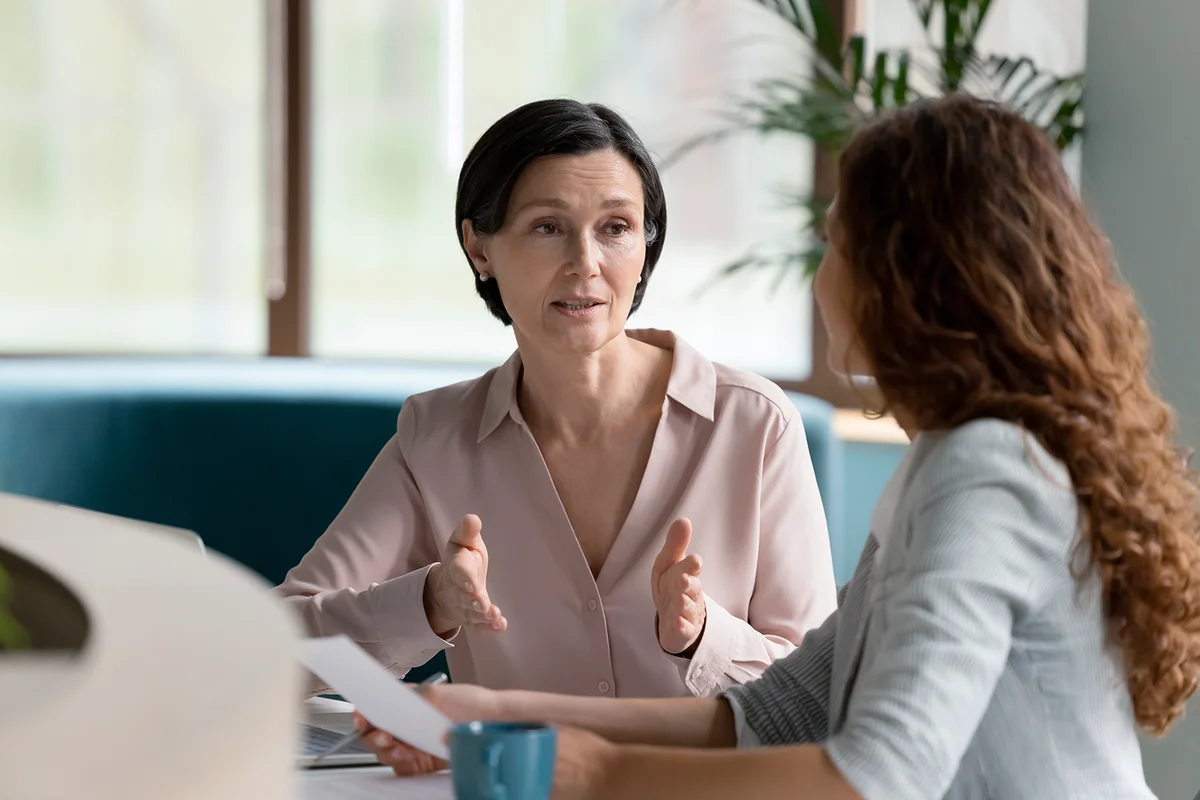 Confident mature business professional woman talking to younger female colleague at office table