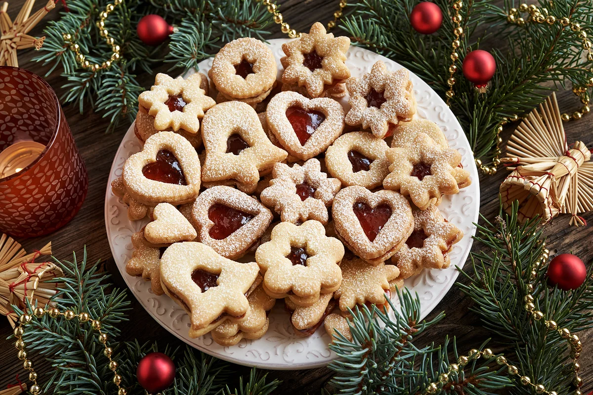A plate of Linzer Christmas cookies filled with strawberry jam and dusted with sugar, with spruce tree branches and straw ornaments
