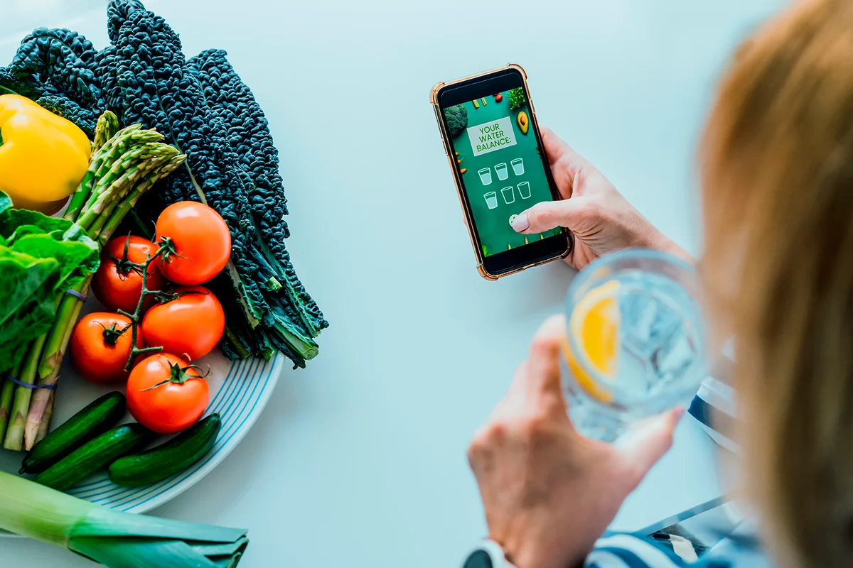 Healthy habit to drink water. Middle age woman drinking glass of pure water with lemon and checking phone while sitting on her kitchen. Using mobile app to control body hydration, Track water balance