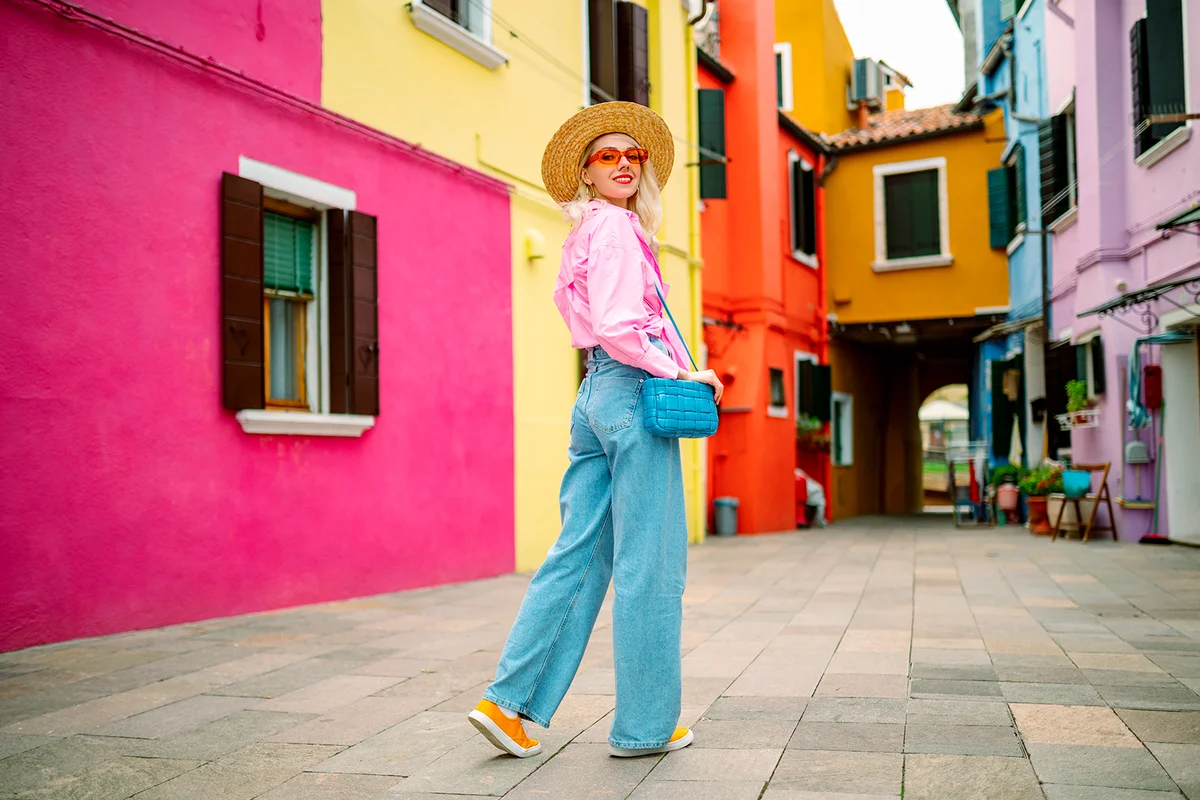 Happy smiling female traveler wearing stylish hat, glasses, pink shirt, wide leg trousers, walking, posing near colorful houses in street. Travel, tourism, vacation, fashion, lifestyle conception