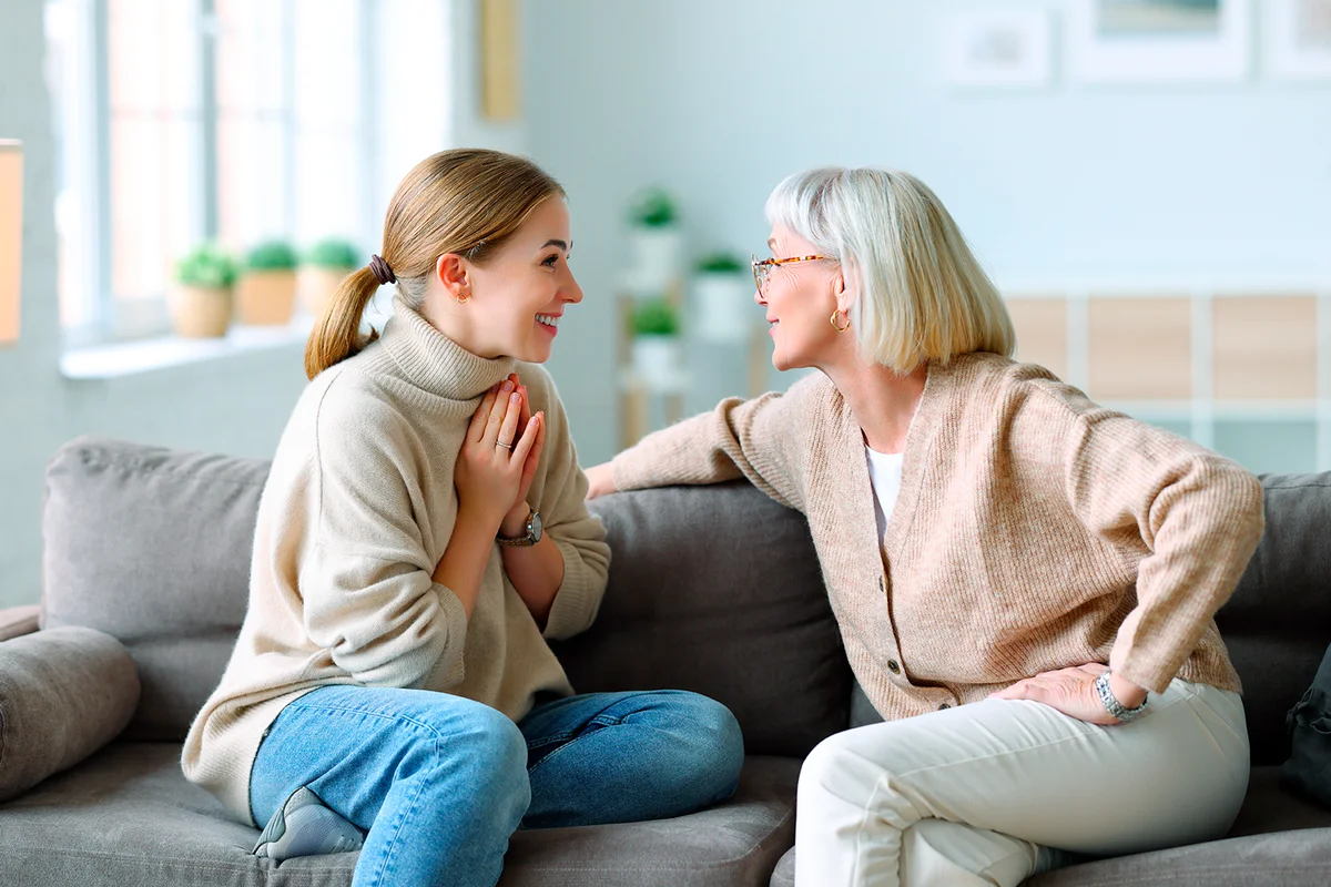 Adult daughter gesticulating and speaking with cheerful laughing elderly mother while resting on couch at home together