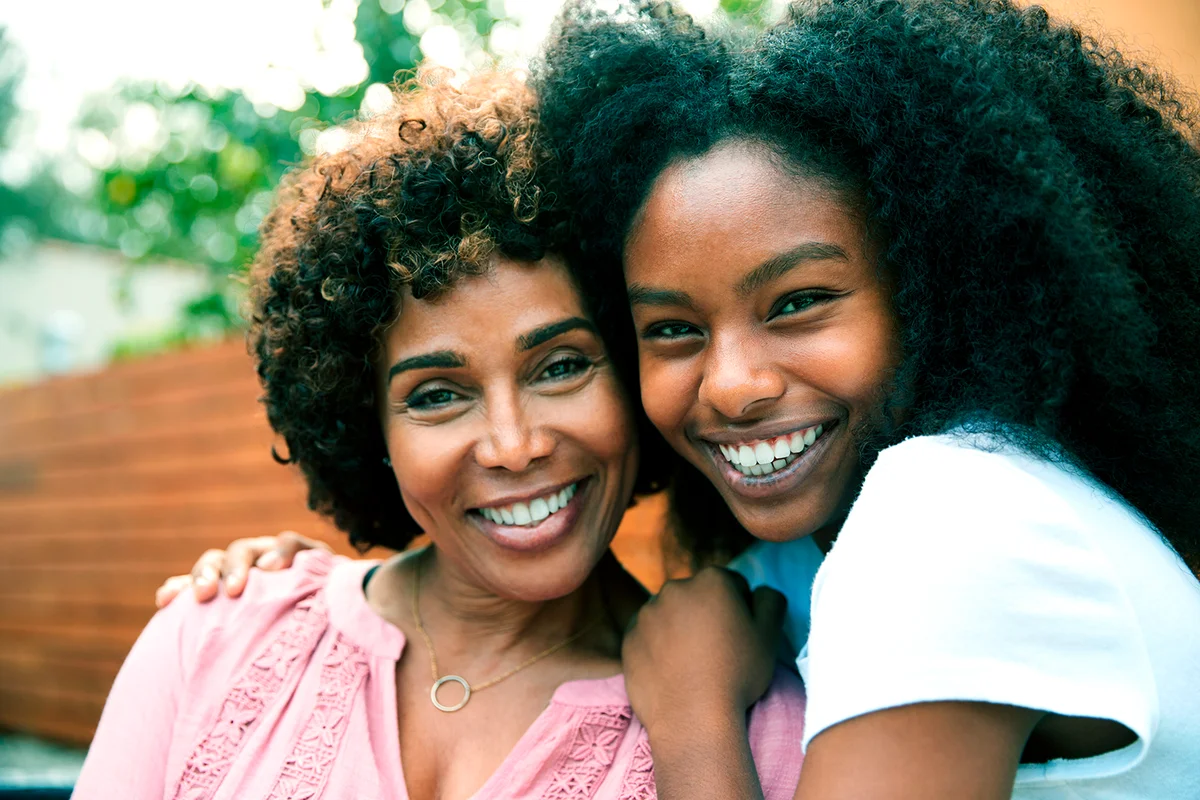 Middle aged black mum and teenage daughter embracing and smiling to camera