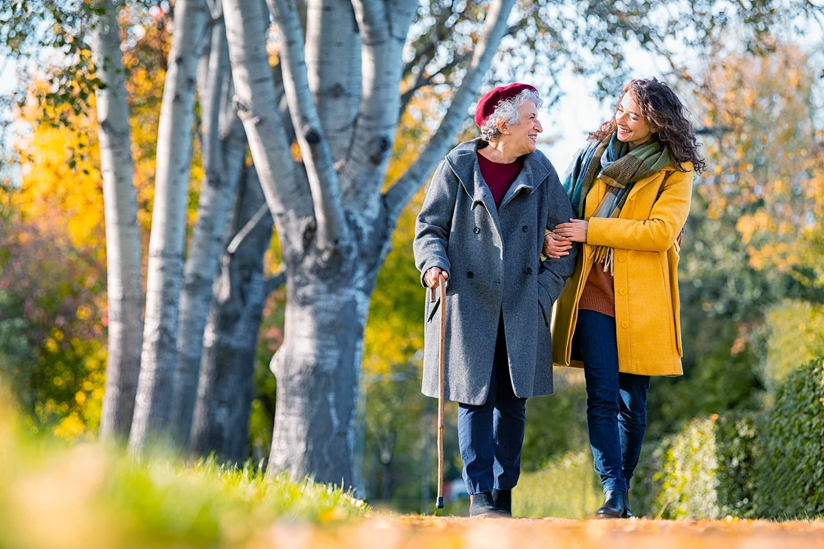 Granddaughter walking with senior woman in park wearing winter clothing. Old grandmother with walking cane walking with lovely caregiver girl. Happy woman and smiling grandma walking in autumn park.