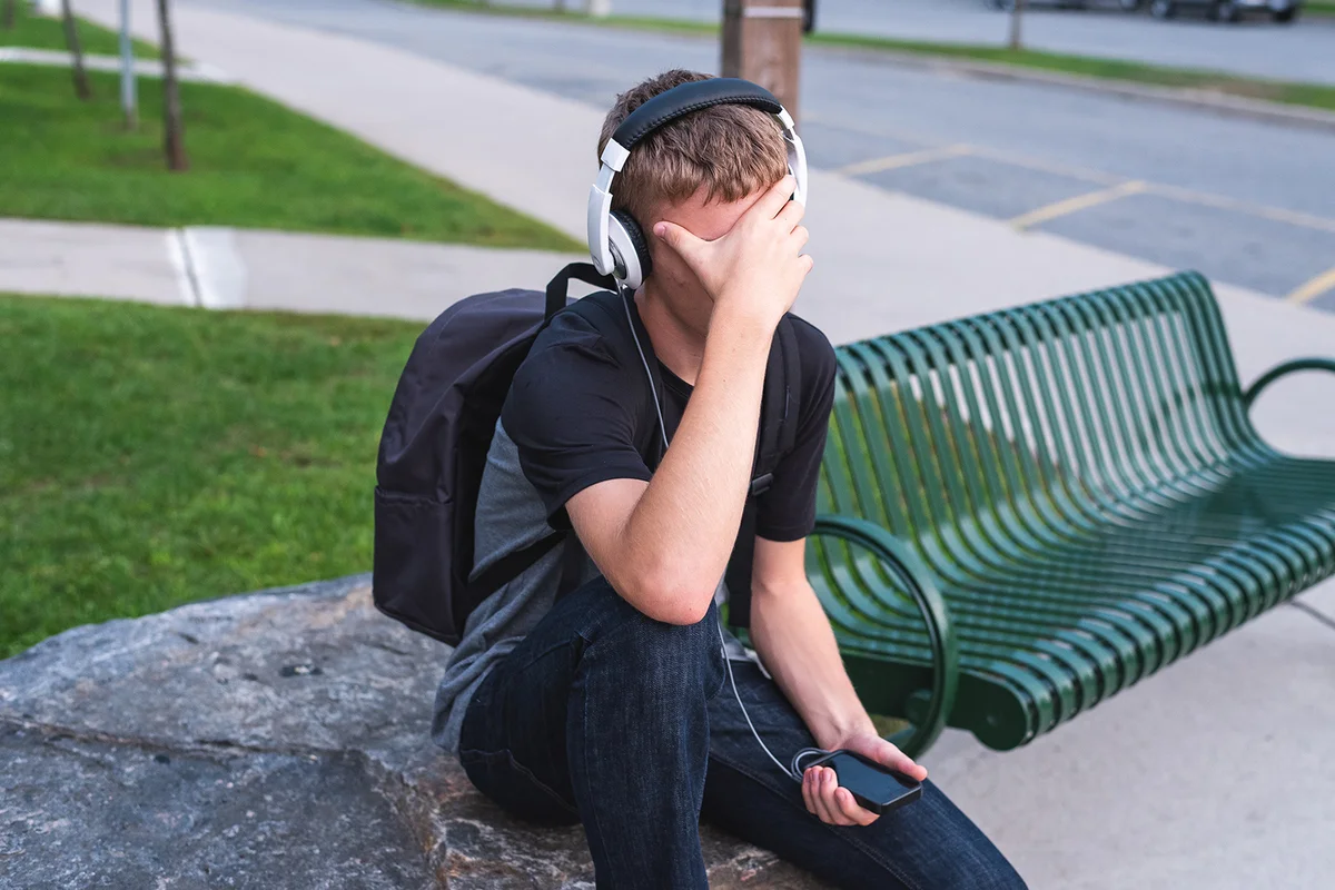 Embarrassed teenager sitting on a rock outside of a school while listening to music