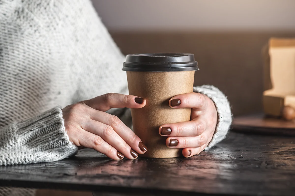 Women's hands in a warm sweater are holding a paper cup of aromatic coffee