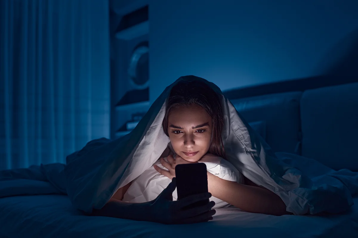 Woman lying under blanket on bed and watching horror movie on mobile phone