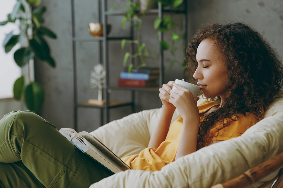 woman drinks coffee reads a book closes her eyes sits in armchair