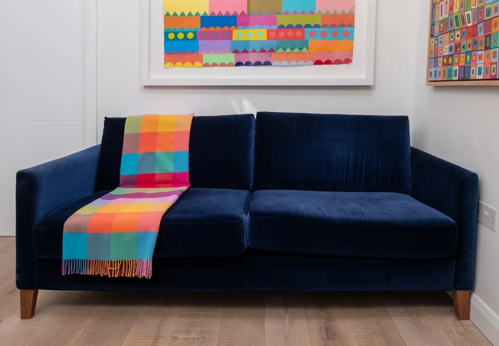 London UK. November 2022. Velvet blue sofa with colourful wool throw, in minimalist setting with white walls and colourful artwork