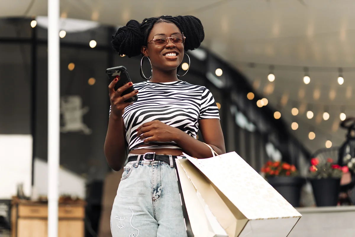 Beautiful fashionable woman holding bags, using smartphone and smiling