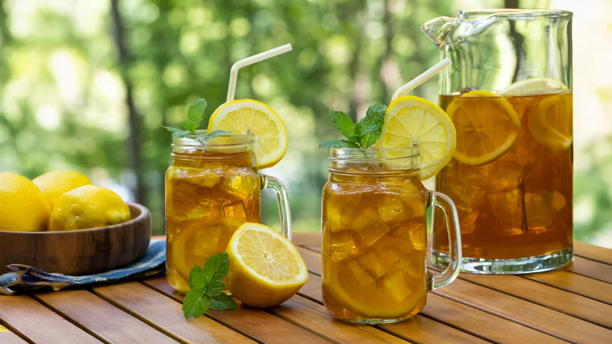 Ice tea in glasses and pitcher with lemon and mint on wooden table with nature background