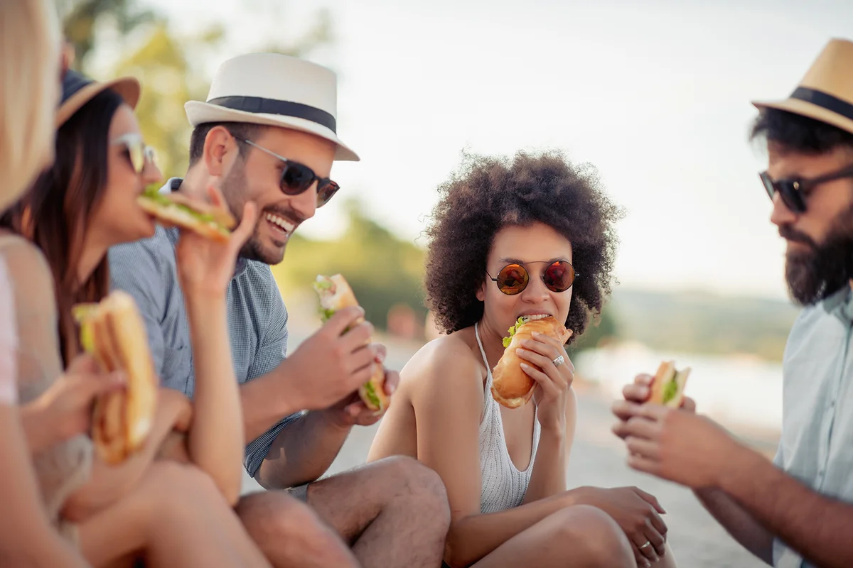 Group of friends sitting on the beach and eating sandwiches during picnic.