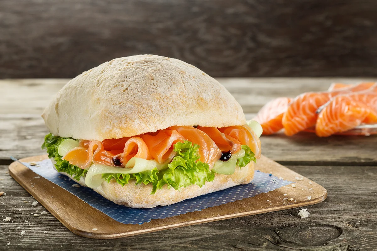 Ciabatta burger sandwich with salmon, cucumber and salad served on the wooden table