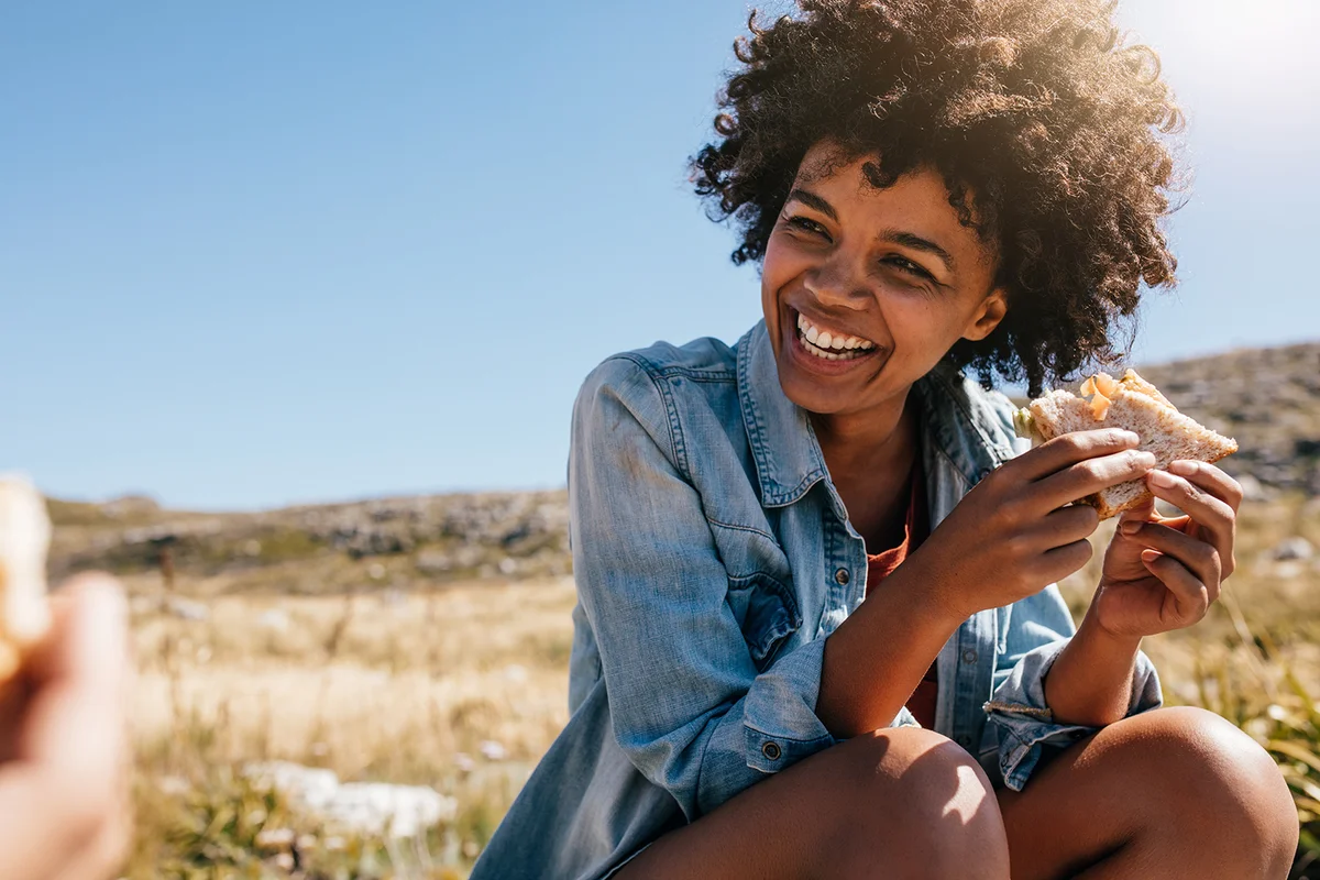 Happy young woman eating sandwich and smiling. Taking break during country hike.