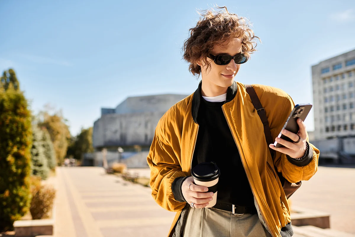A young man in sunglasses checks his phone while sipping coffee in a busy plaza.