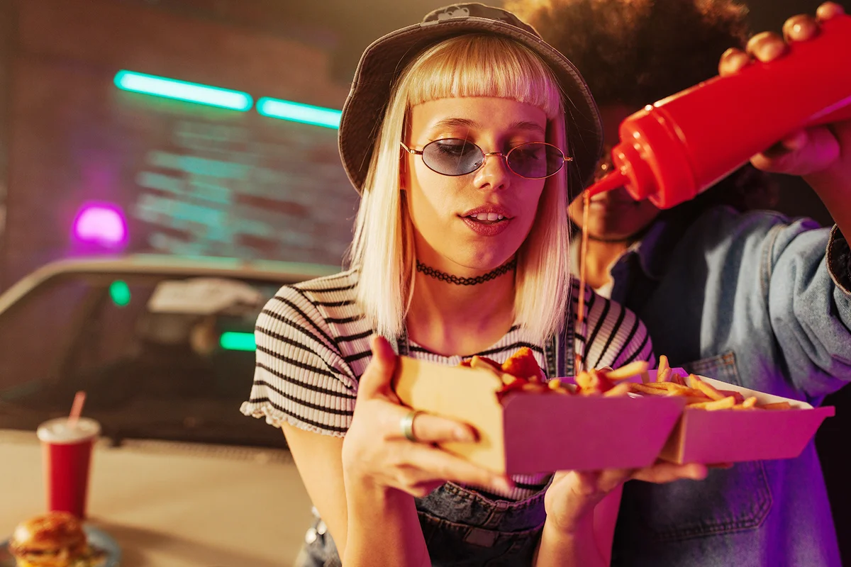 Two stylish girls are using ketchup on their fries while sitting on the hood of their car