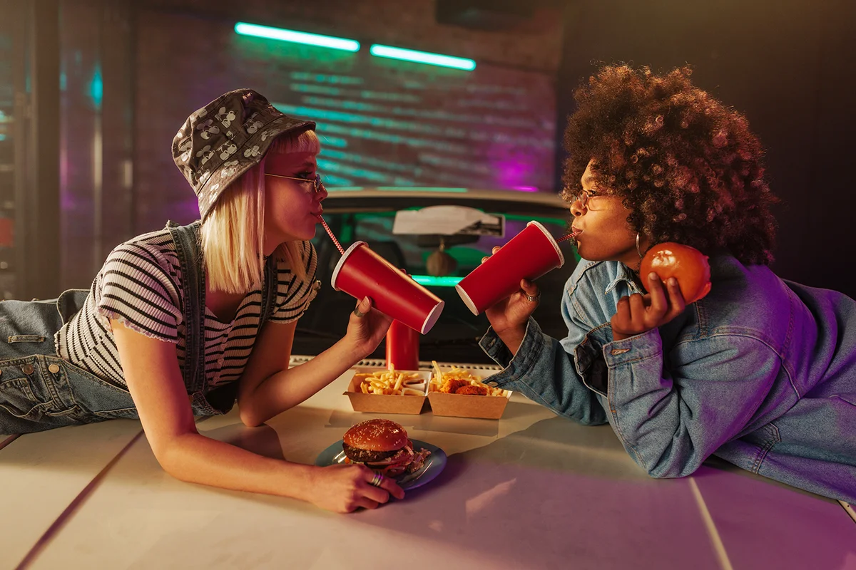 Two young adult fashionable women are eating some takeout food on a hood of their car.