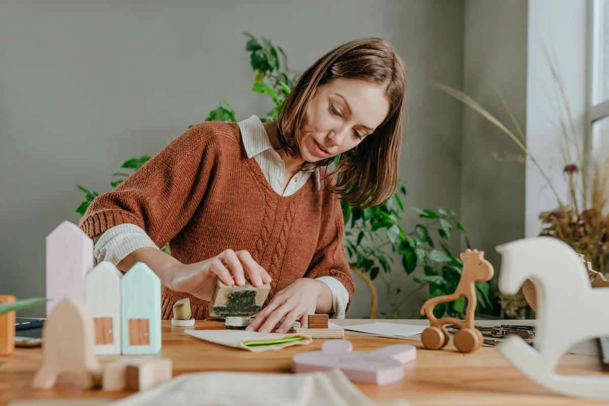 A woman crafts at a table surrounded by wooden supplies using a rubber stamp.