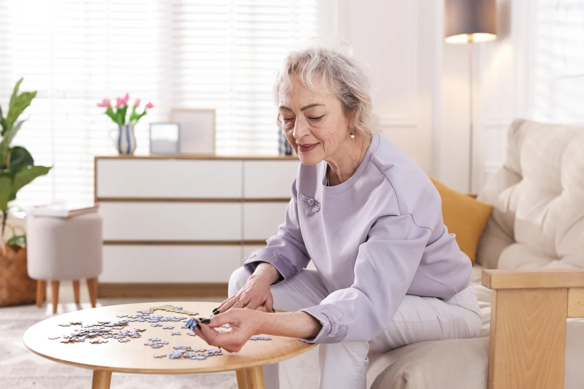 An elderly woman works on a jigsaw puzzle at a small table.